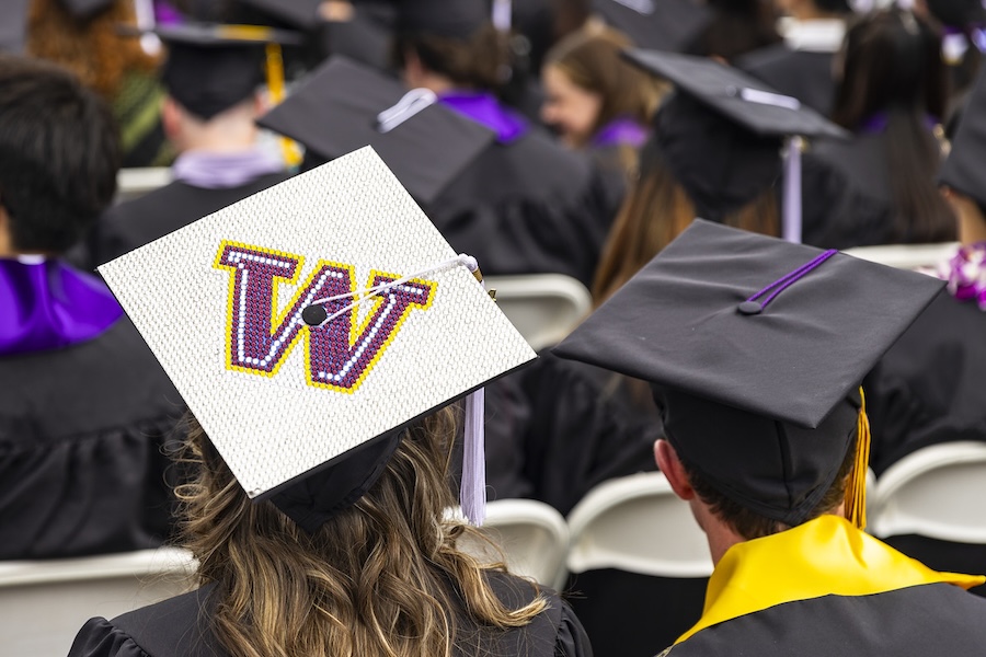 View from behind a crowd of graduating students wearing graduation caps. Features a graduation cap/mortarboard decorated with a purple W on a white background.