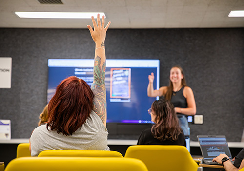 A person raises their hand in a classroom in the foreground and the teacher is pointing at them