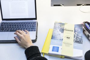 Closeup of a desk showing someone's hand using a laptop and some books.